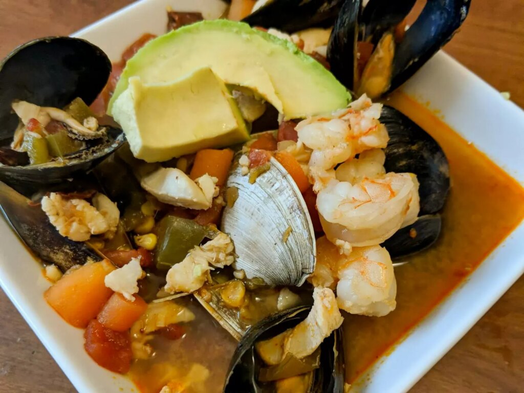 Close up view of sopa de mariscos (seafood soup) with clams, muscles, shrimp, carrots, peppers and avocado, in a white bowl