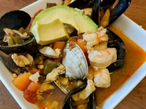 Close up view of sopa de mariscos (seafood soup) with clams, muscles, shrimp, carrots, peppers and avocado, in a white bowl