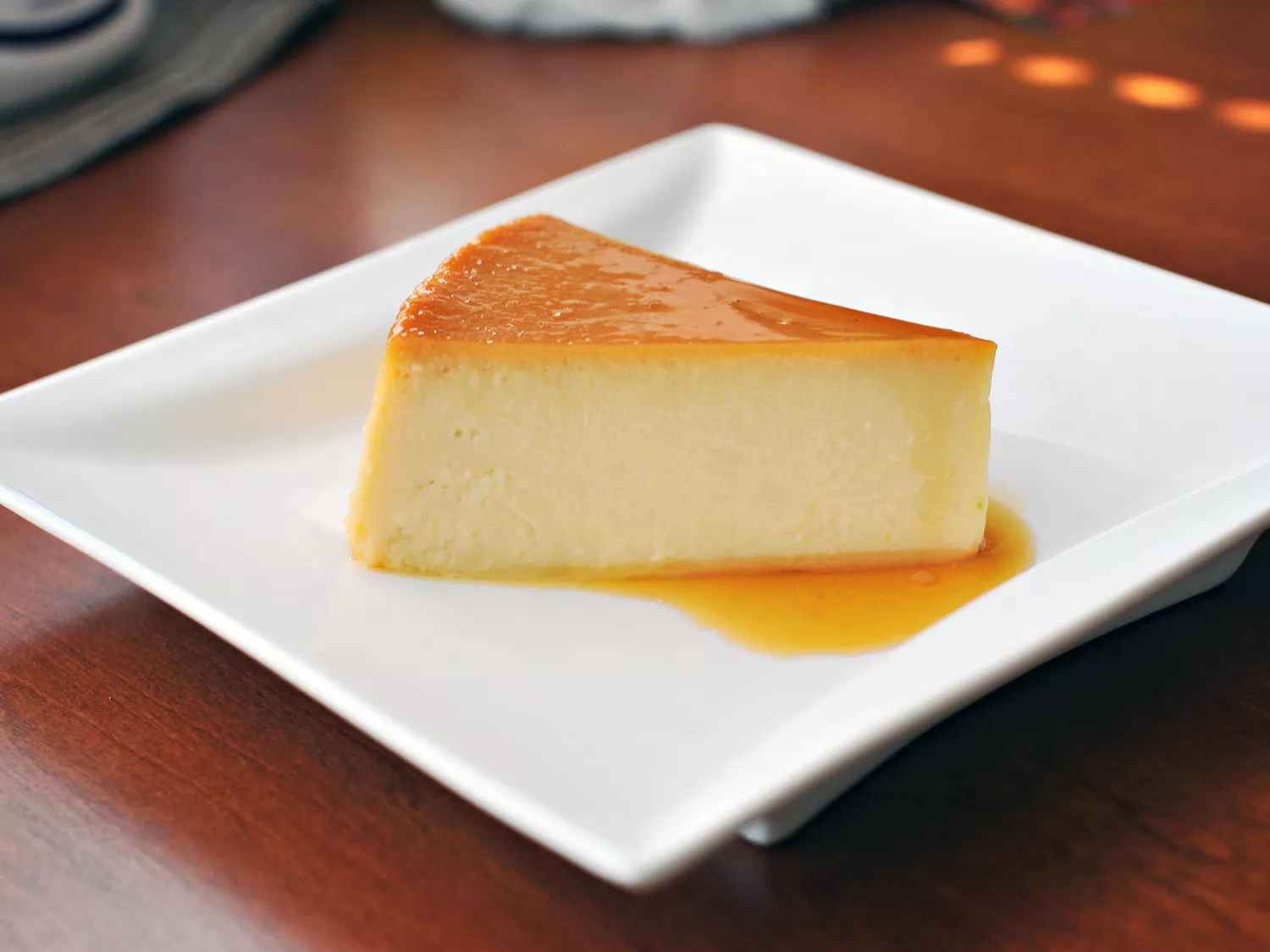 A slice of perfect looking spanish flan with a caramel top layer and pool of sauce on a square white plate