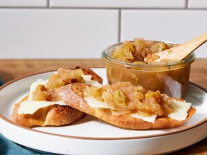 Slices of bread topped with cheese and apple chutney served on a plate with a jar of chutney and wooden spoon beside it