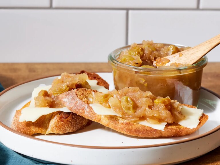 Slices of bread topped with cheese and apple chutney served on a plate with a jar of chutney and wooden spoon beside it