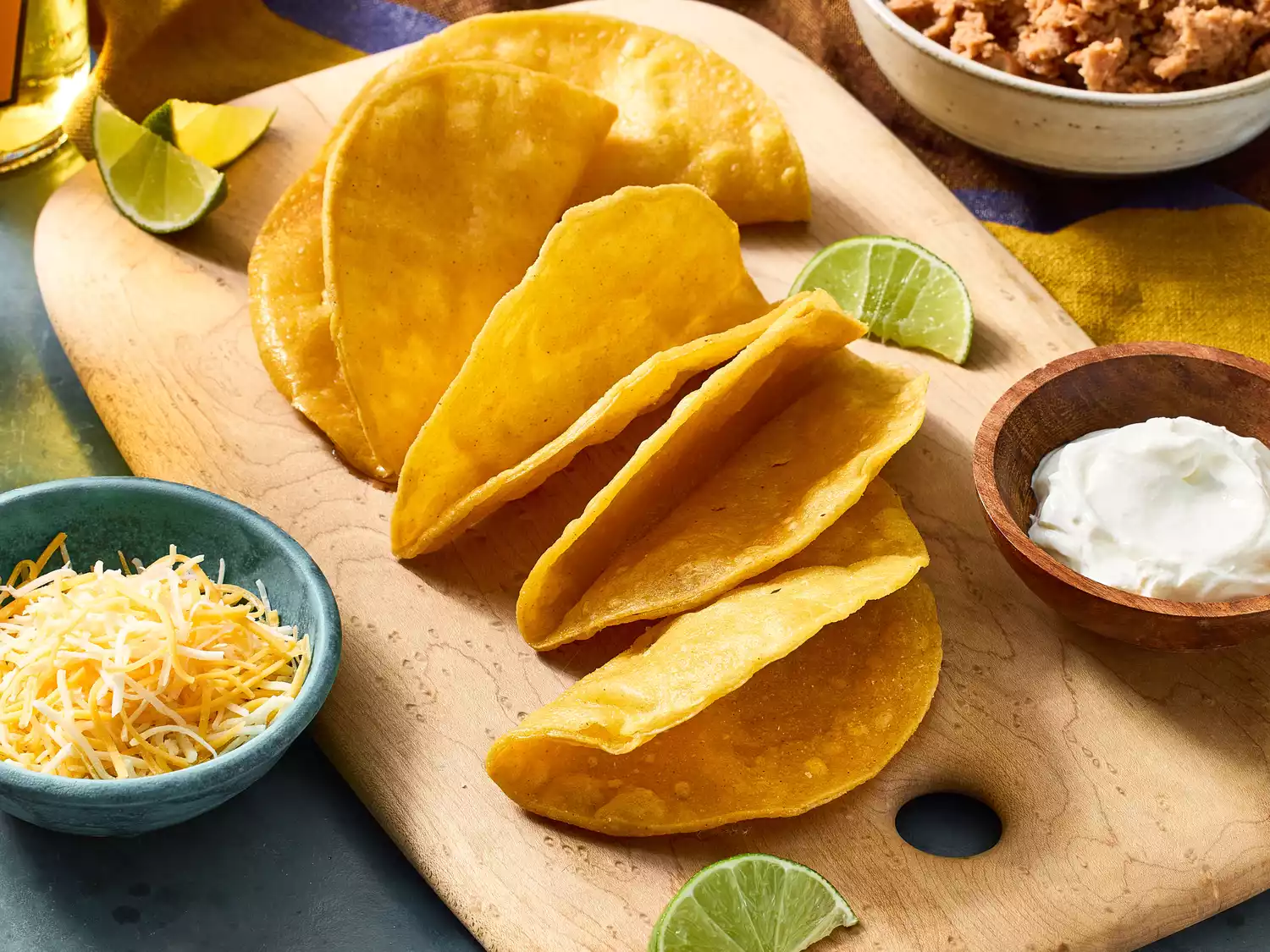Crispy taco shells on wooden board with cheese, sour cream, and lime.
