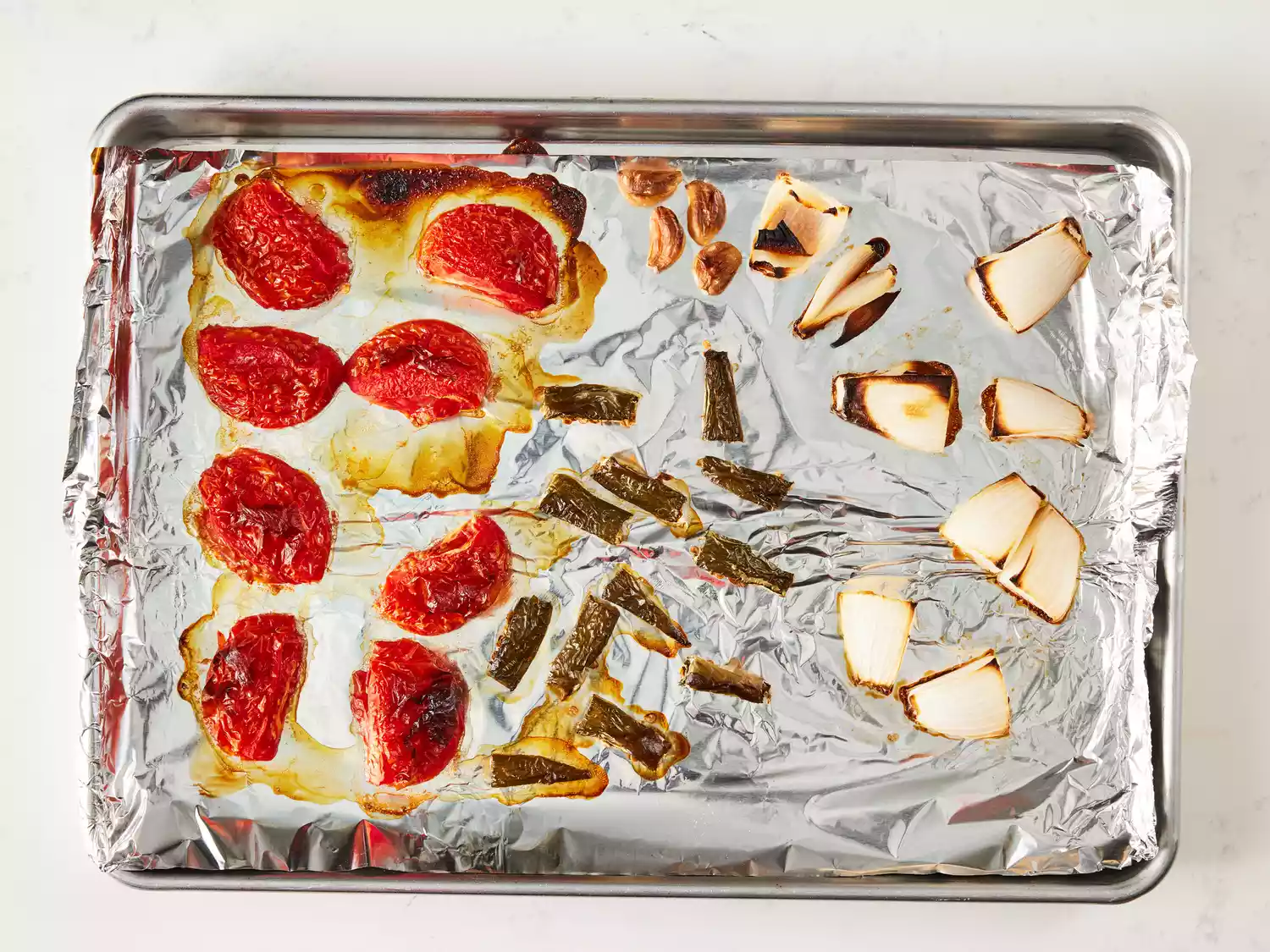 an overhead shot of roasted tomatoes, onions, cloves of garlic, and jalapeÃ±os on a baking sheet