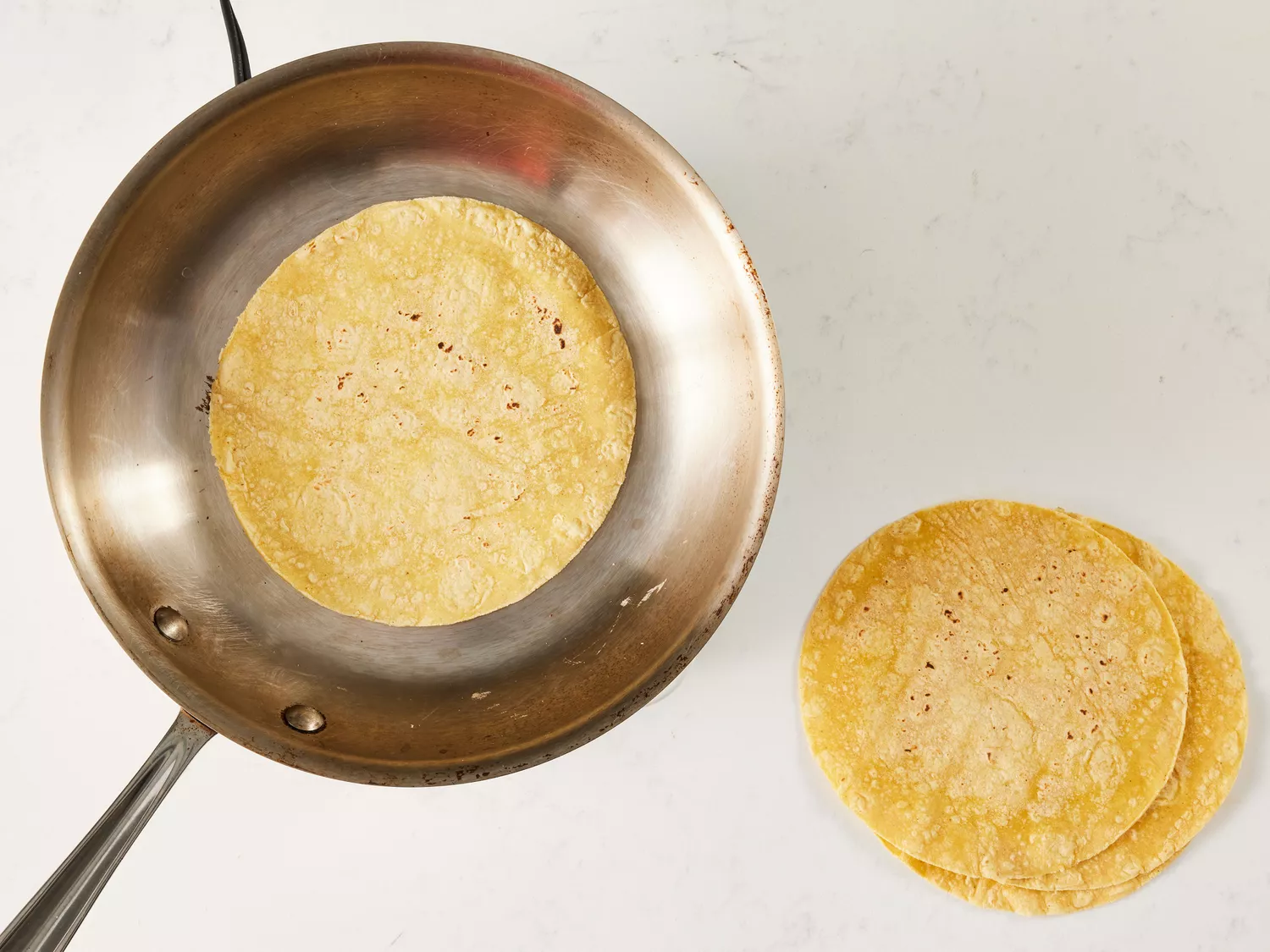 an overhead shot of a tortilla cooking in a skillet and more tortillas on the side