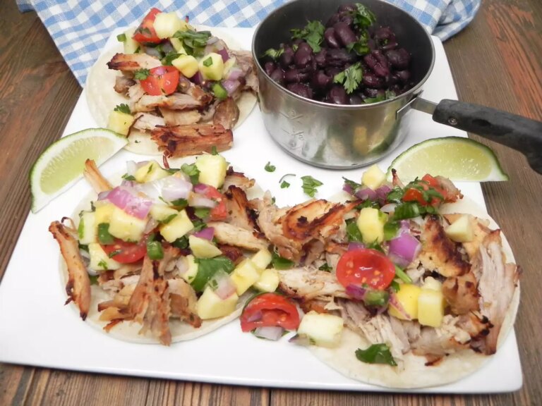 Three street tacos with carnitas, salsa, lime wedges, and pot of black beans on white platter