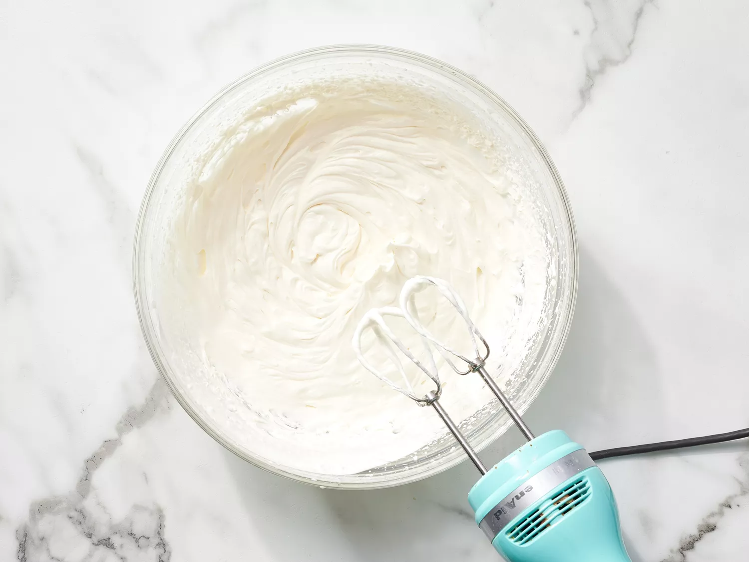 Whipped cream in a glass bowl next to a hand mixer 