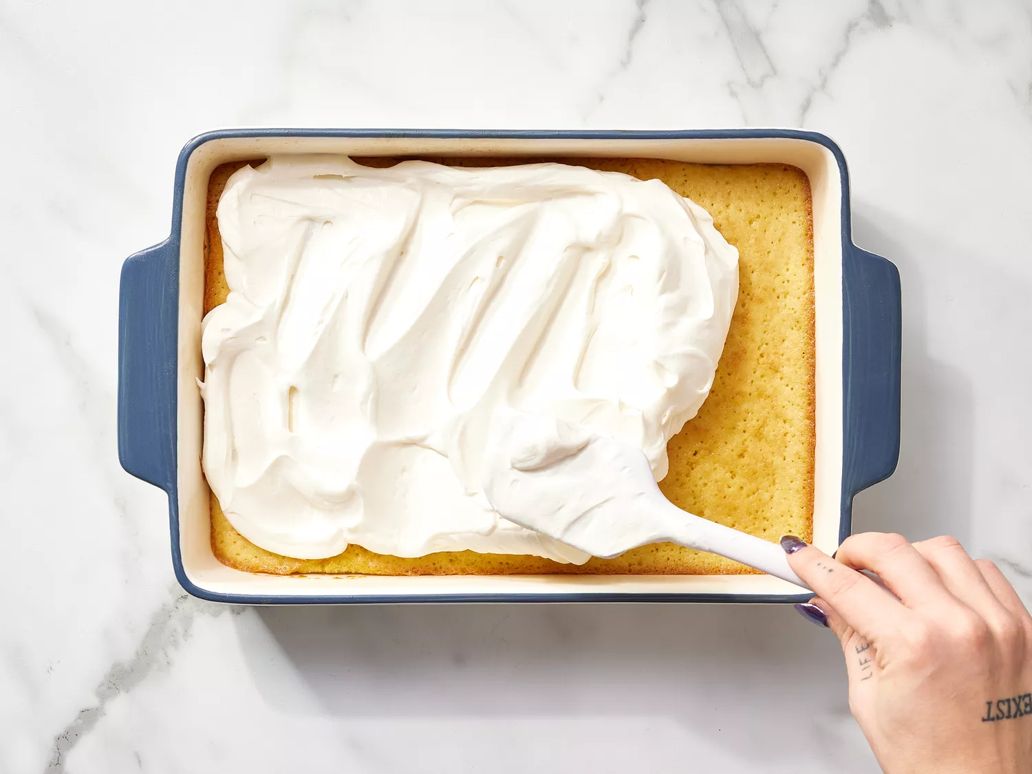 Spatula spreading whipped cream on top of a cake in a baking dish 