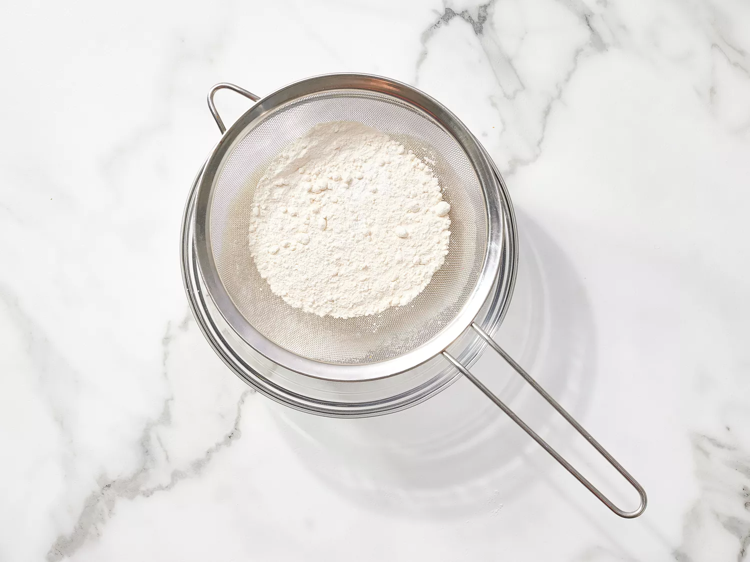 Flour and baking powder in a fine mesh sifter over a glass bowl 