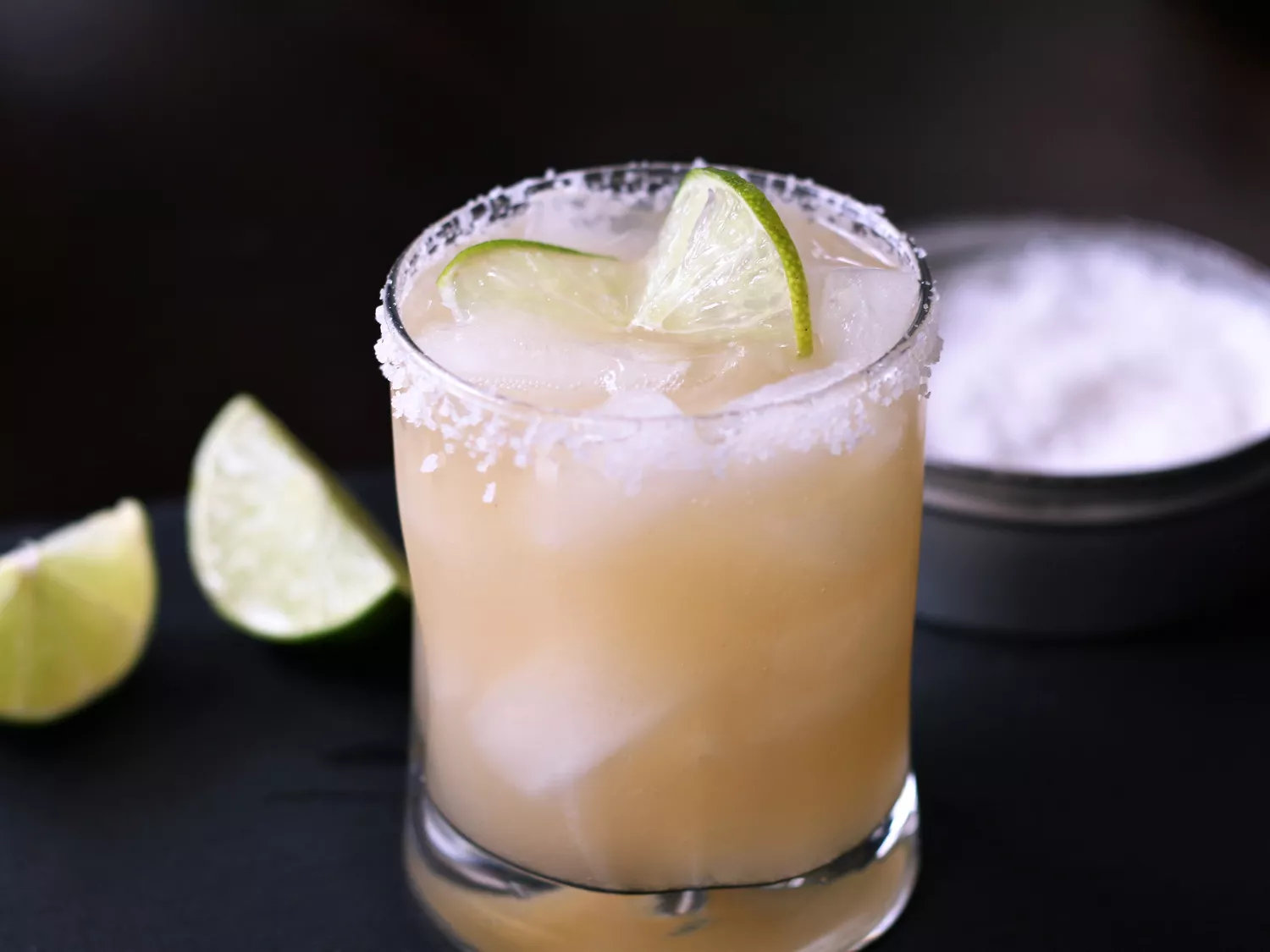 Salt rimmed glass with pale drink on ice with lime wedges on black background