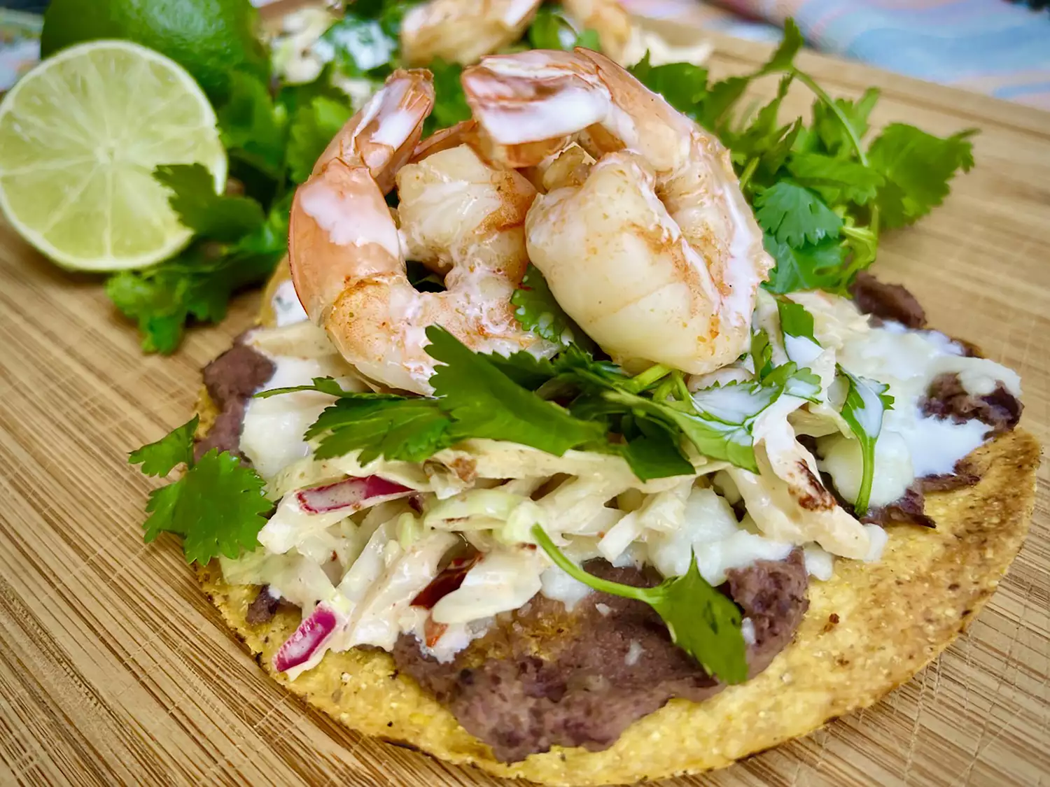 Tostada shell on board with beans, slaw, shrimp, and cilantro with lime in background