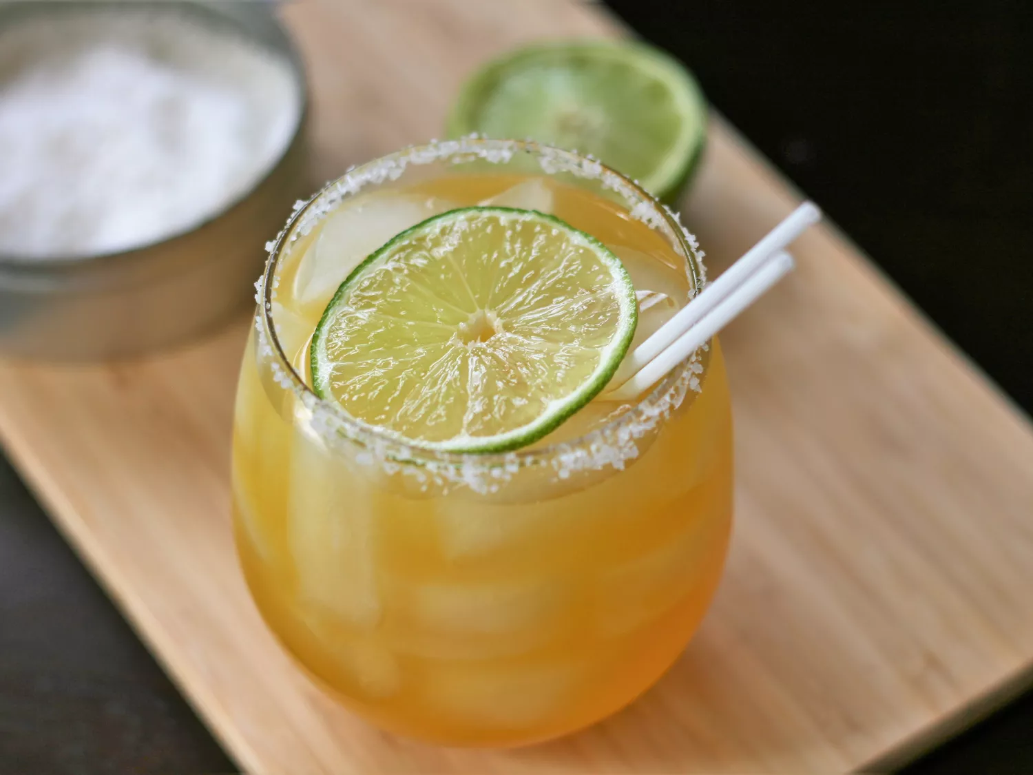Closeup of cocktail with salted rim, lime slice, straws, on a board