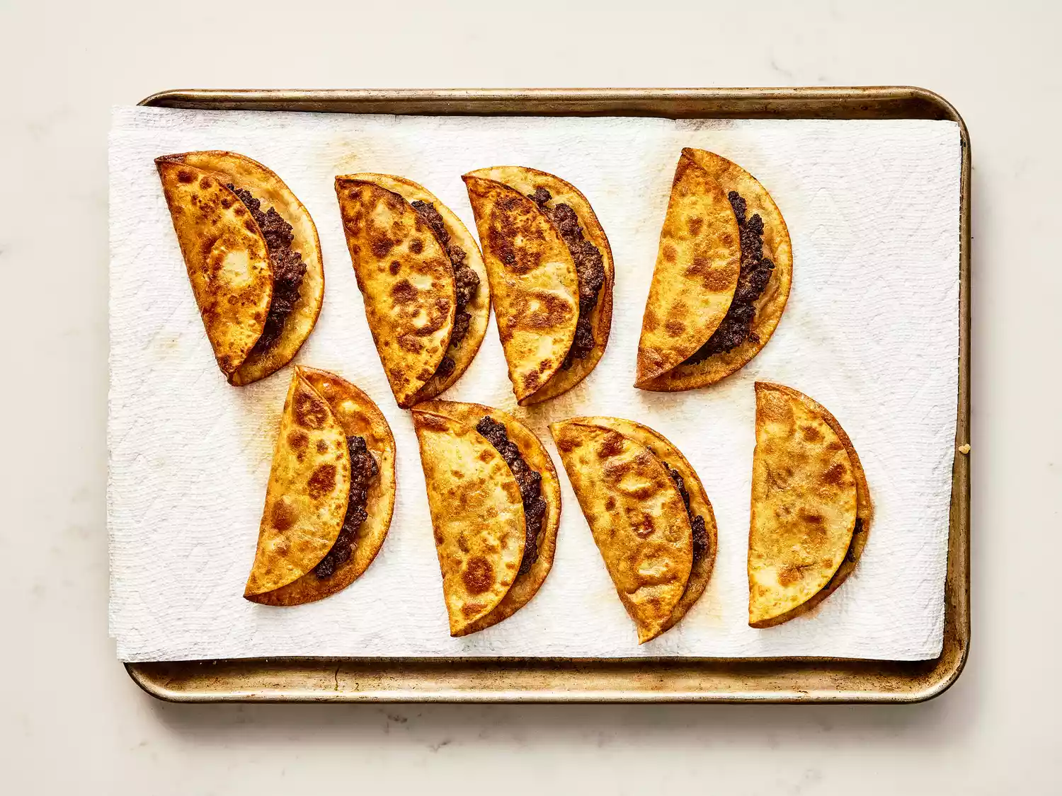8 golden-brown fried beef tacos resting/draining on a paper towel lined baking sheet. 