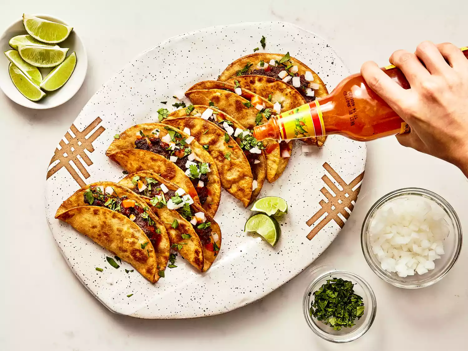8 fried beef tacos on a platter being dressed with onions, cilantro, and hot sauce. 