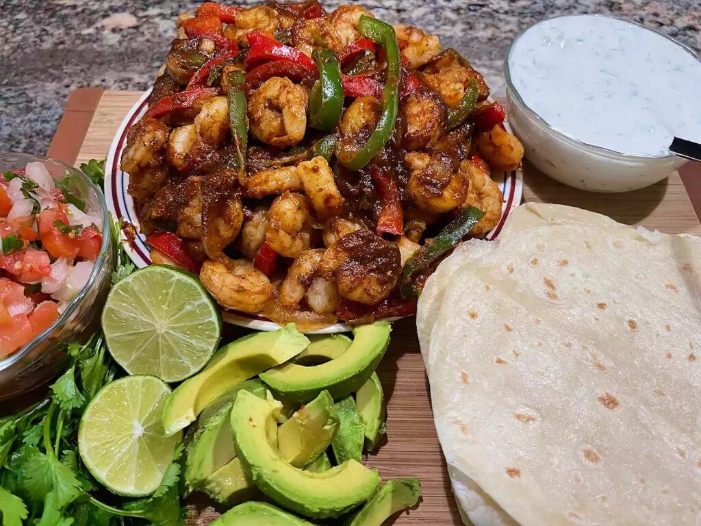 Plate of shrimp and bell pepper fajitas with tortillas, sliced avocado, pico de gallo, and cream sauce on a board