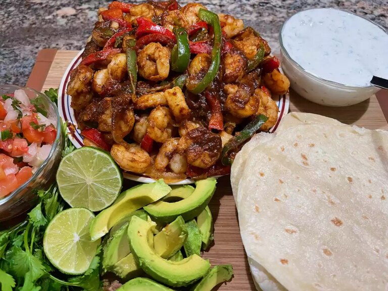 Plate of shrimp and bell pepper fajitas with tortillas, sliced avocado, pico de gallo, and cream sauce on a board
