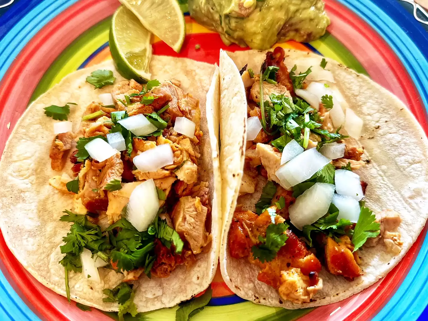 Overhead view of two chicken tacos on a colorful plate with limes and guacamole