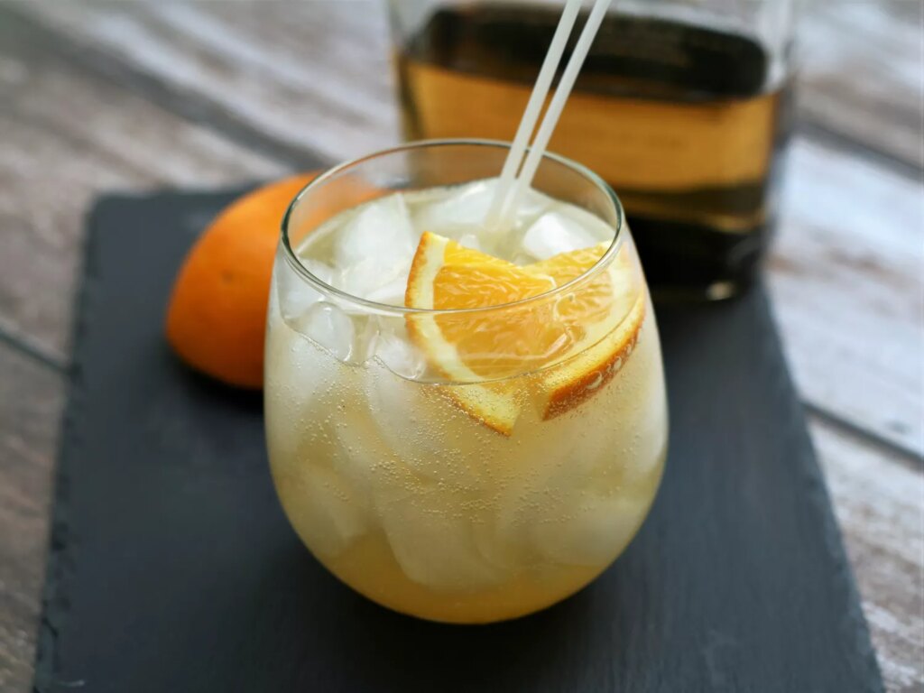 Rocks glass with iced fizzy drink of golden tequila, orange slices, with orange and tequila bottle in background