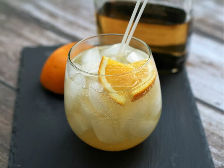 Rocks glass with iced fizzy drink of golden tequila, orange slices, with orange and tequila bottle in background