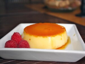 Closeup side view of an individual serving of flan inverted onto a white square plate so the amber caramel layer is on top, with three fresh raspberries on the side