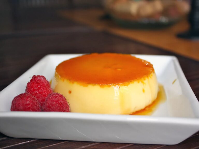 Closeup side view of an individual serving of flan inverted onto a white square plate so the amber caramel layer is on top, with three fresh raspberries on the side