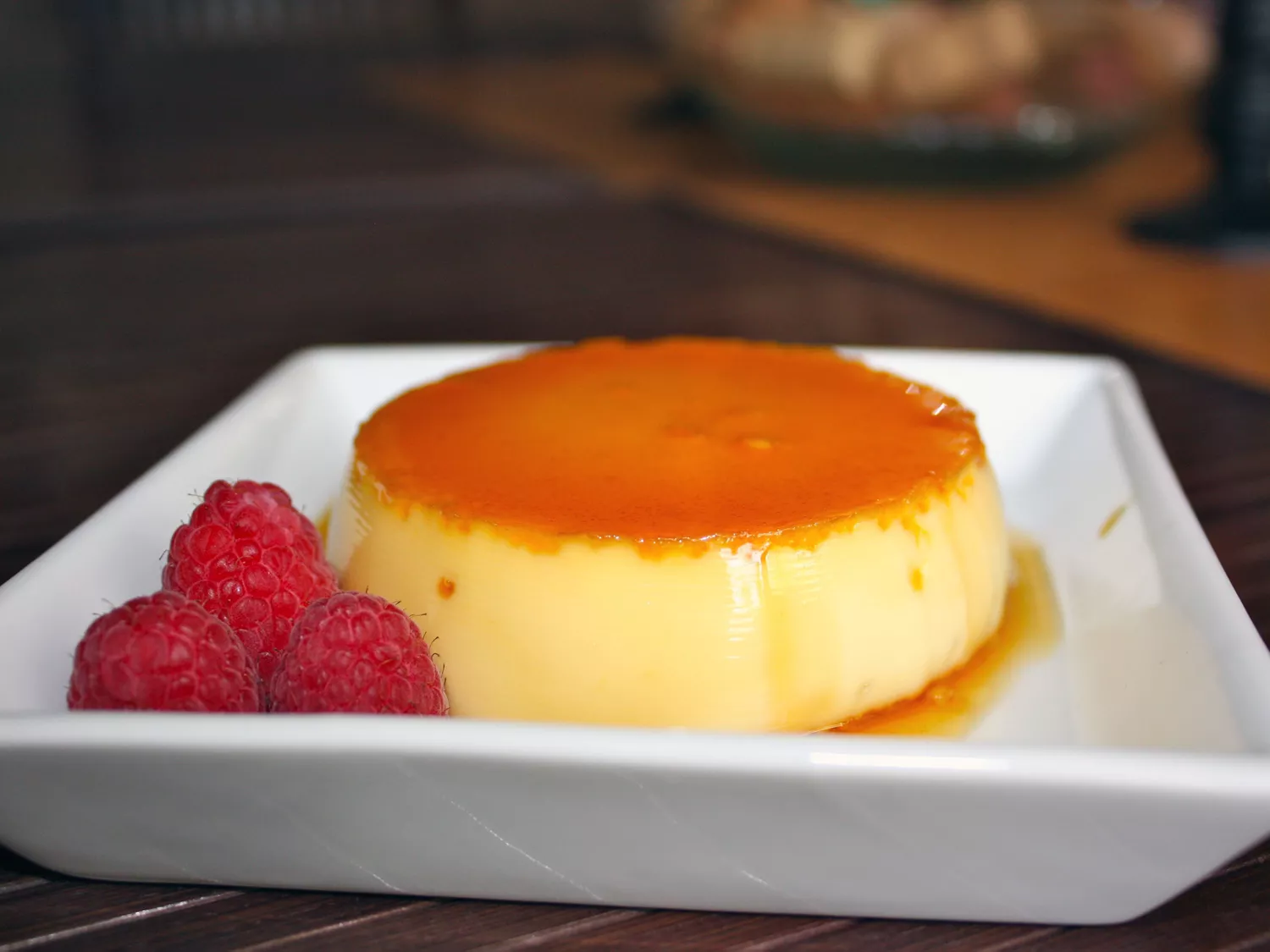 Closeup side view of an individual serving of flan inverted onto a white square plate so the amber caramel layer is on top, with three fresh raspberries on the side