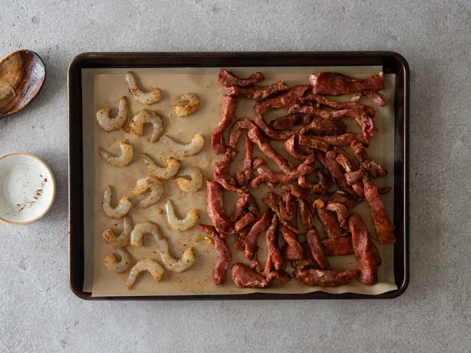 A baking sheet containing seasoned raw shrimp and steak strips placed side by side prepared for cooking