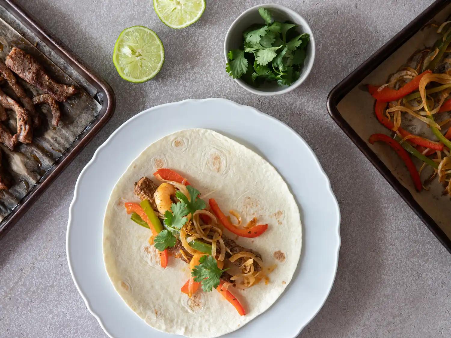 A tortilla with steak shrimp and fajita vegetables on a plate surrounded by cooking trays and garnishes
