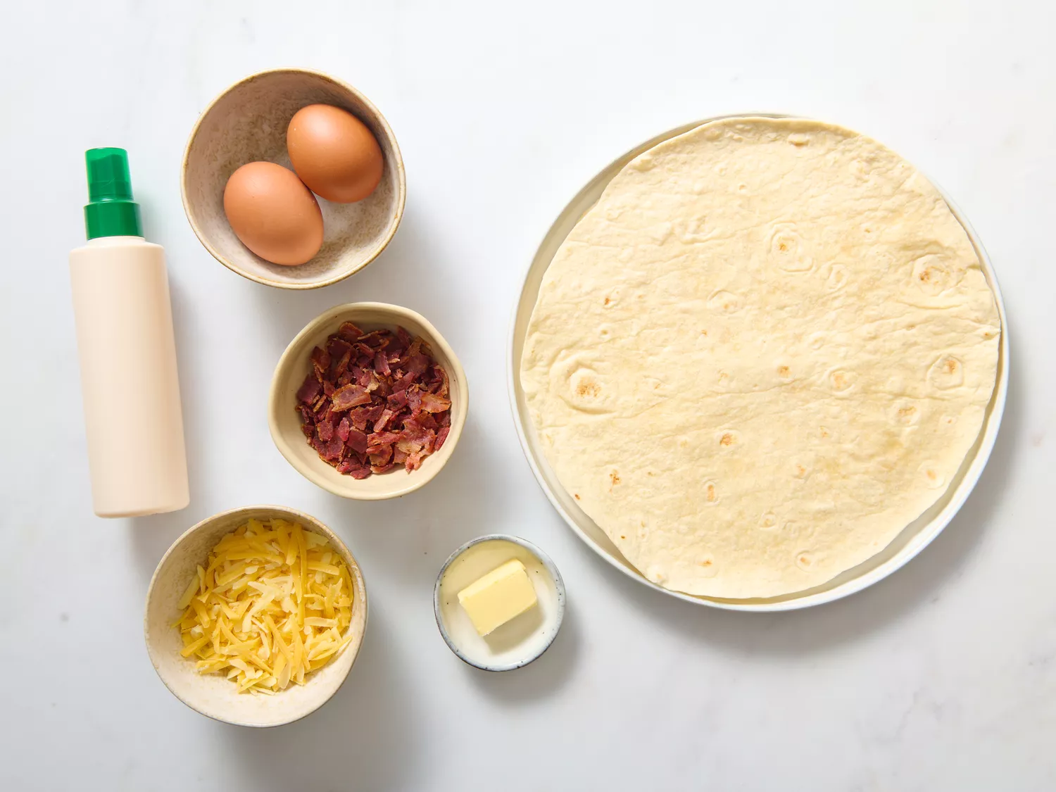 Ingredients for making breakfast quesadillas on a table, including tortillas, eggs, shredded cheese, cooked bacon, butter, and sauce bottle