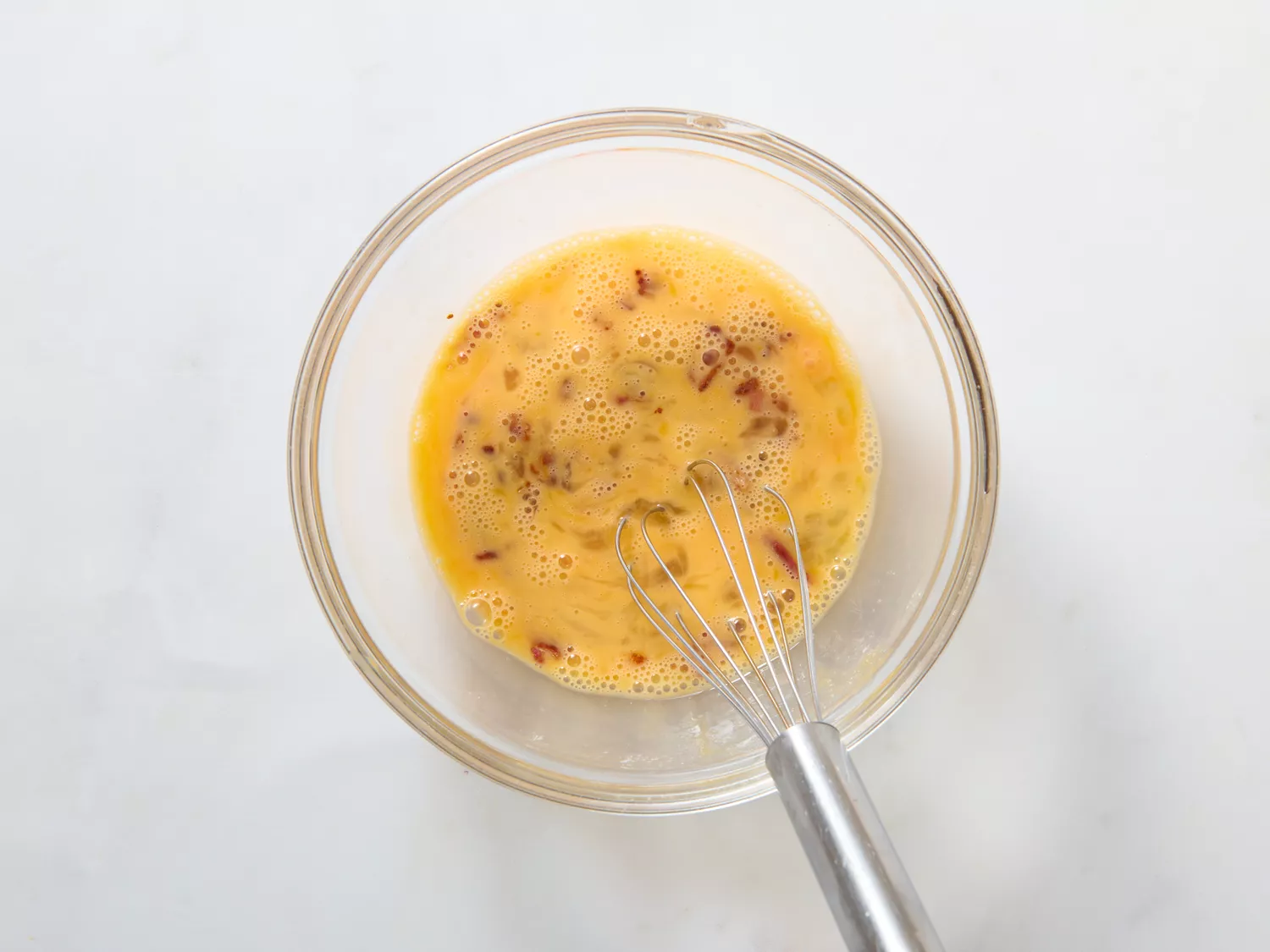 A bowl of whisked eggs with small bits of cooked bacon, a whisk placed inside