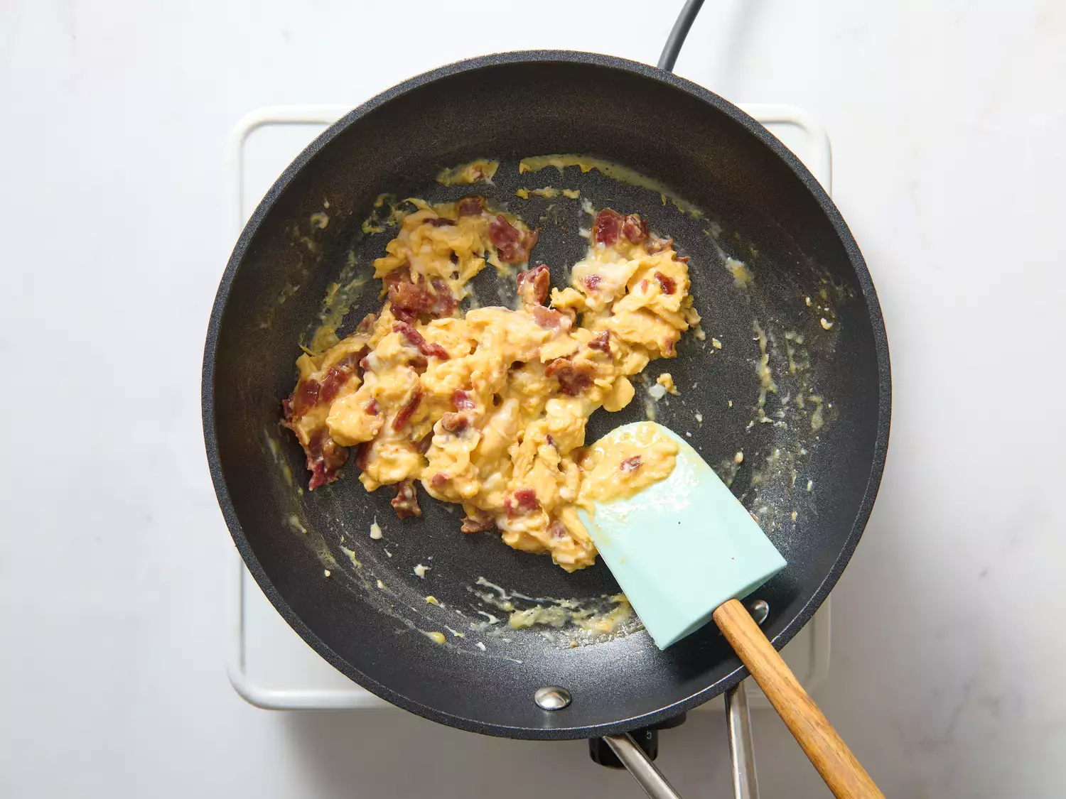Scrambled eggs with pieces of cooked bacon in a frying pan with a spatula