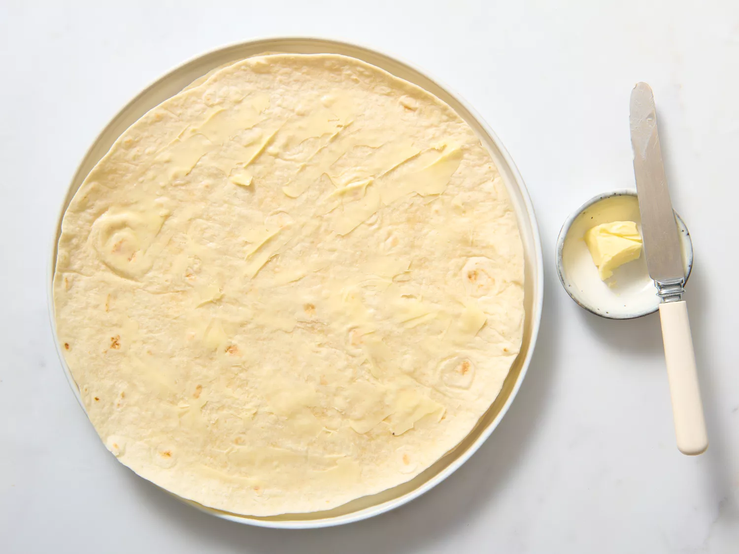 A tortilla being spread with butter on a plate, beside a butter knife and a small dish of butter