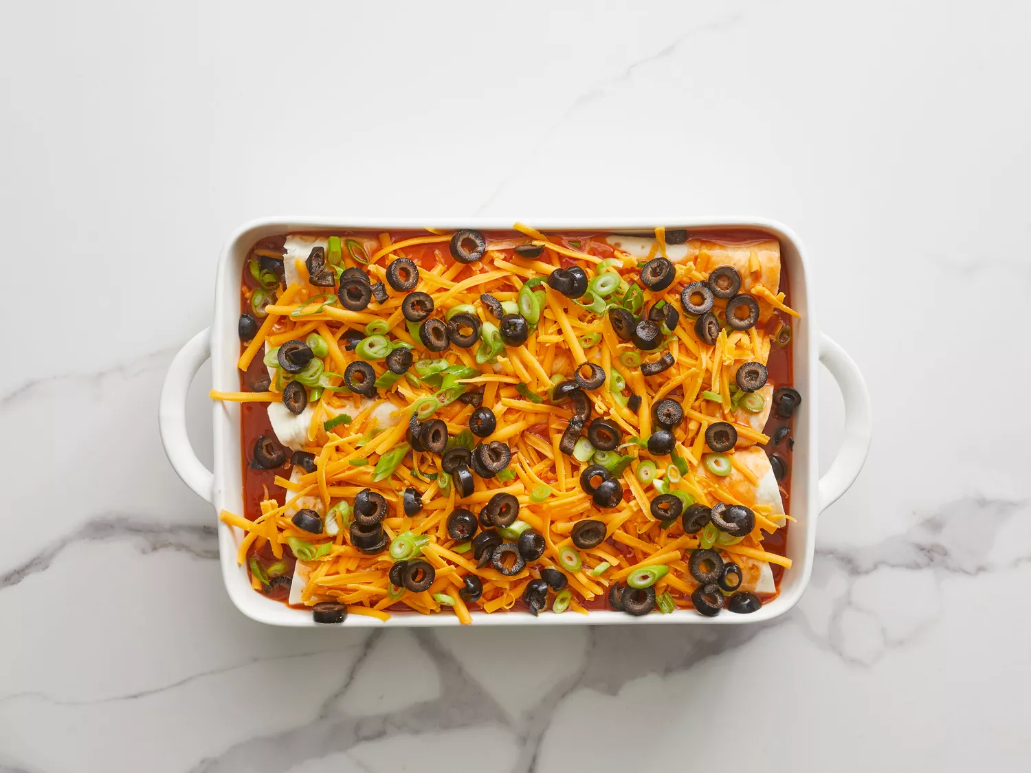 an overhead shot of enchiladas topped with sauce, cheddar cheese, olives, and green onions in a baking dish