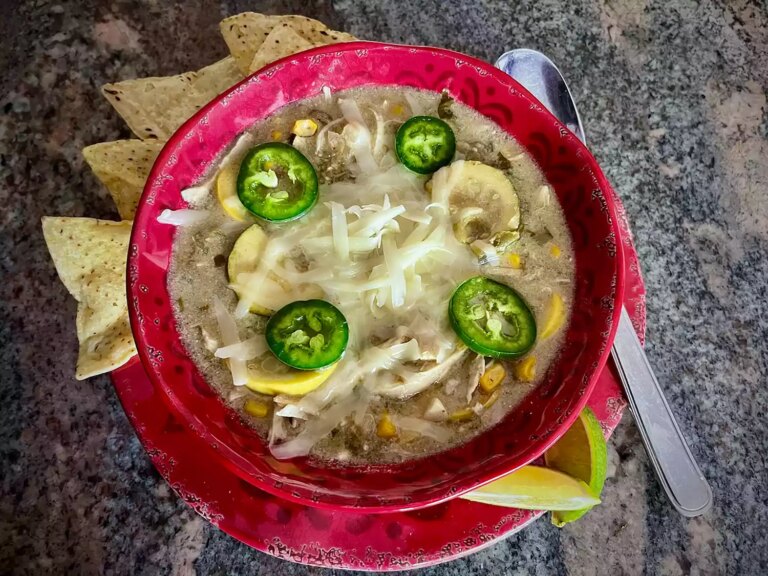 Red soup bowl with green chicken enchilada soup, topped with jalapeno slices and grated cheese, with lime slices and tortilla chips on the side