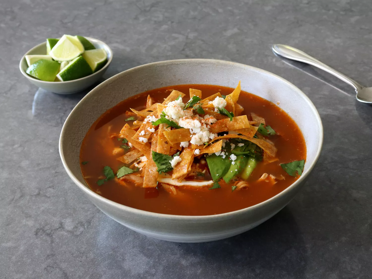 White bowl of chicken tortilla soup with deep red broth, tortilla strips, avocado slices, and crumbled cheese