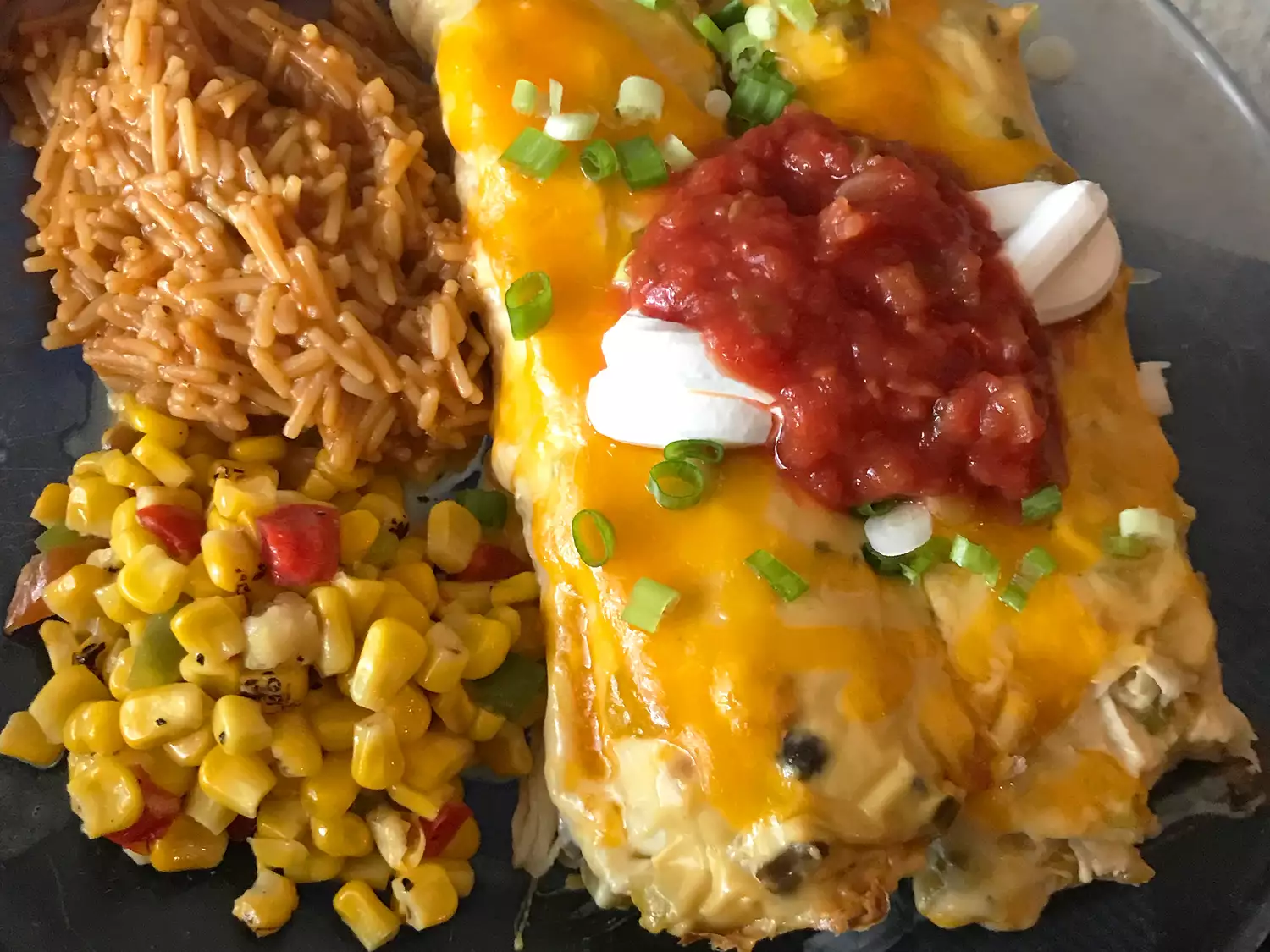 Close up view of chicken enchiladas v topped with green onions, salsa and sour cream, with rice and corn on the side