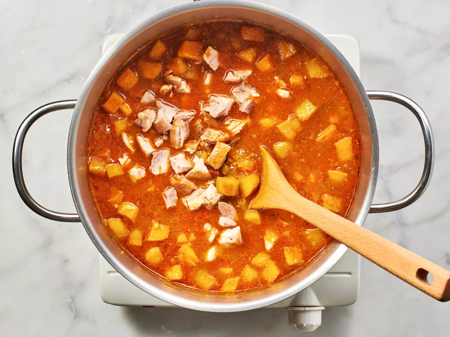 A wood spoon in a pot of soup with diced chicken and potatoes.