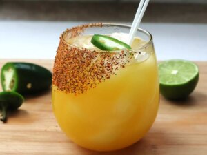 Stemless wine glass with mango margarita, salted with tajin, limes and jalapenos in background