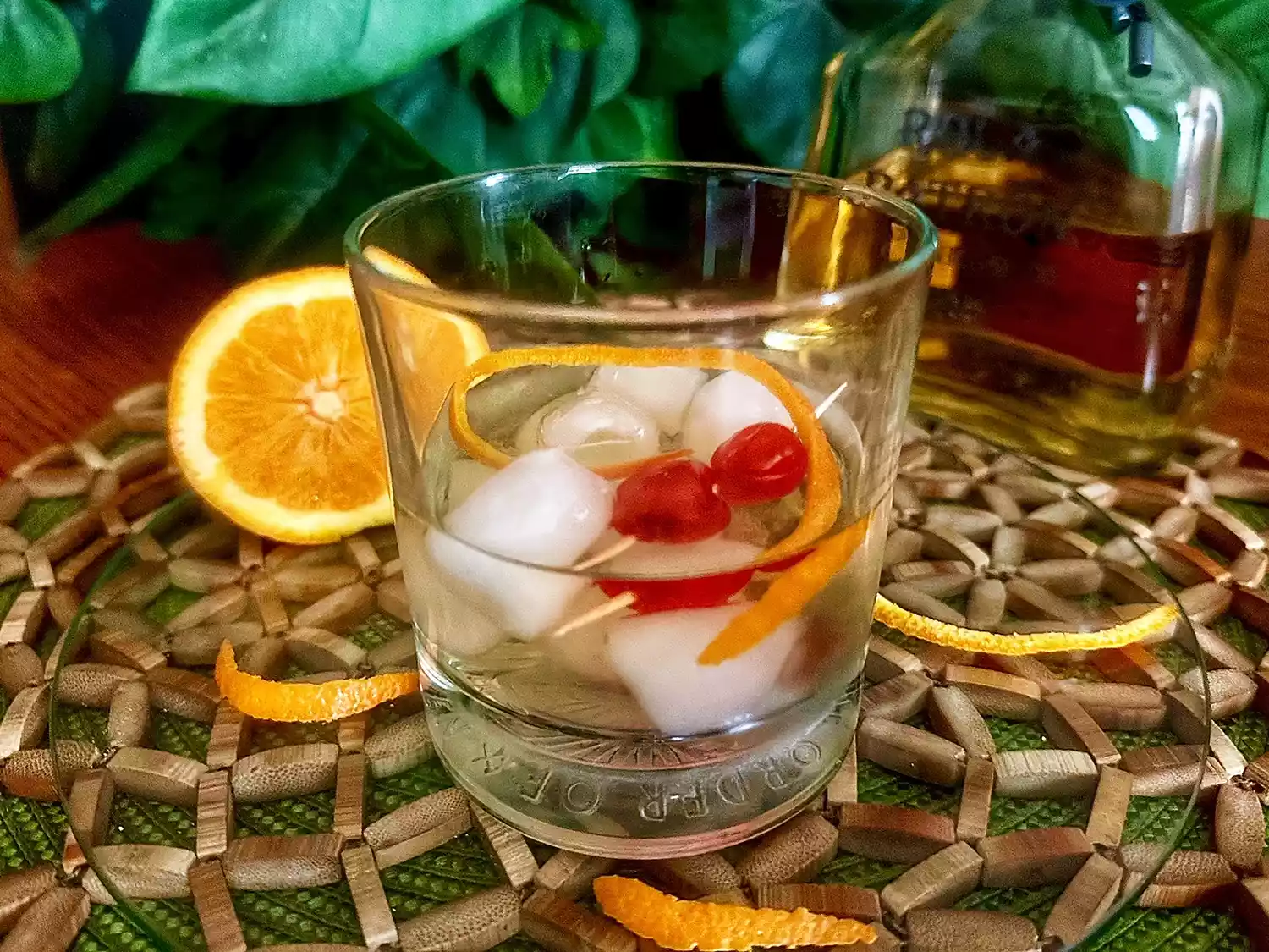 Rocks glass with old fashioned and cherries on woven beaded mat with orange half in background