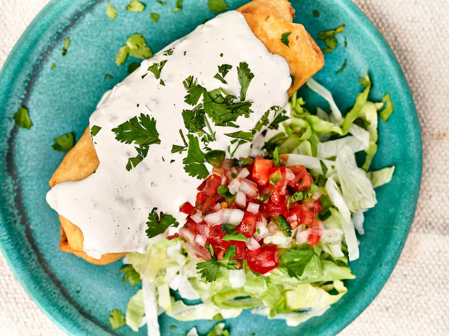 Overhead view of chimichanga on teal plate with crema, lettuce, and pico de gallo