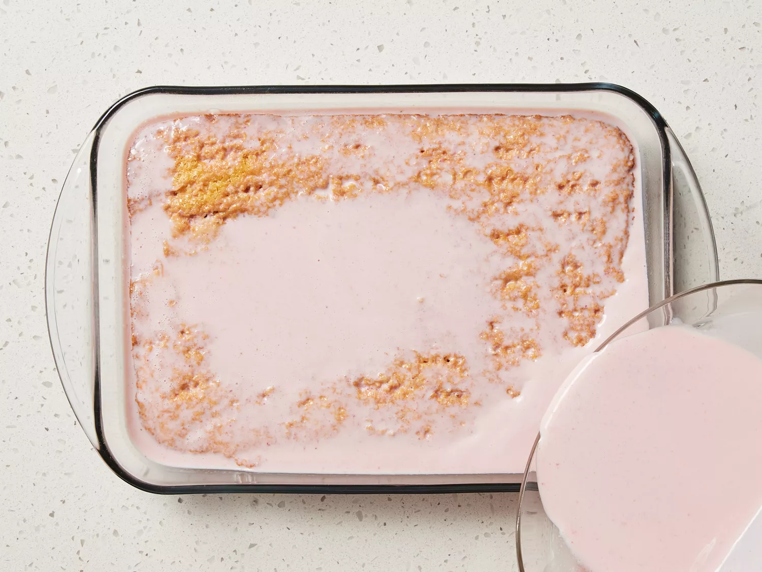 Strawberry milk being poured over a cake in a glass baking dish 