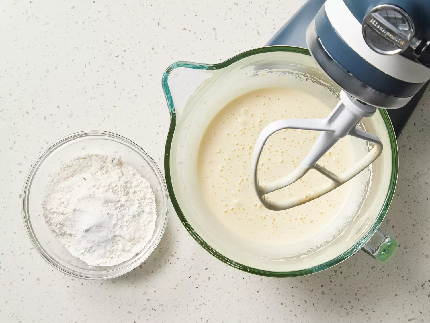 Batter in the glass bowl of a stand mixer next to flour in a glass bowl 