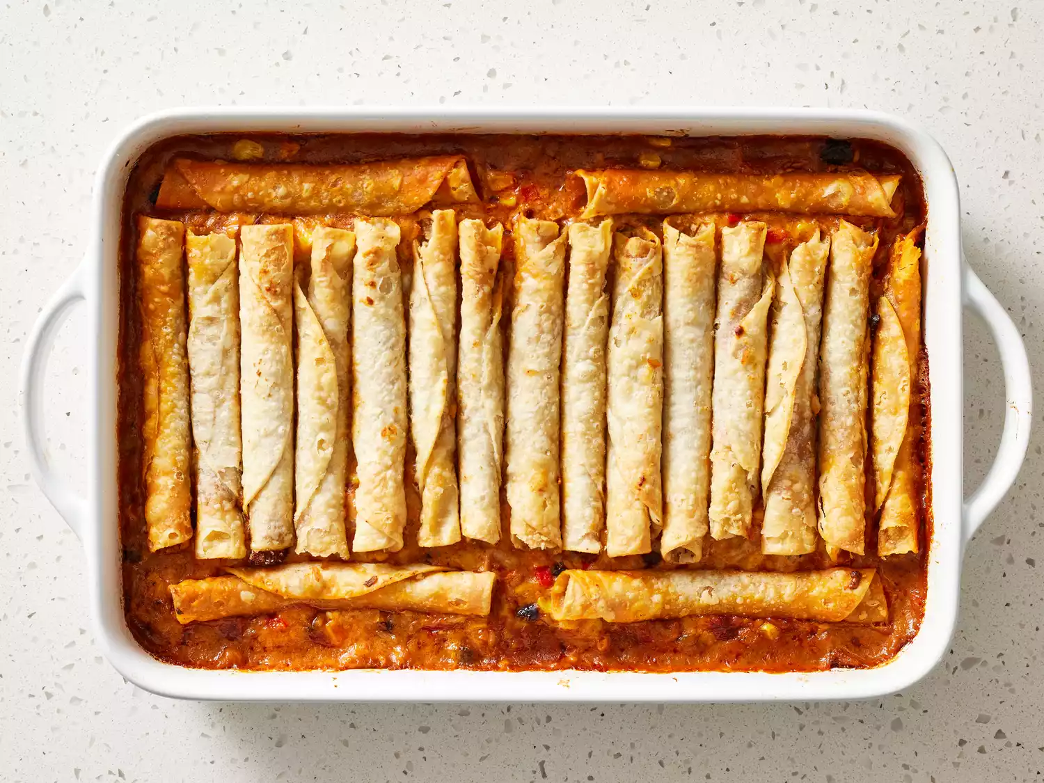 Taquitos on top of cheese and red sauce mixture in a white baking dish 
