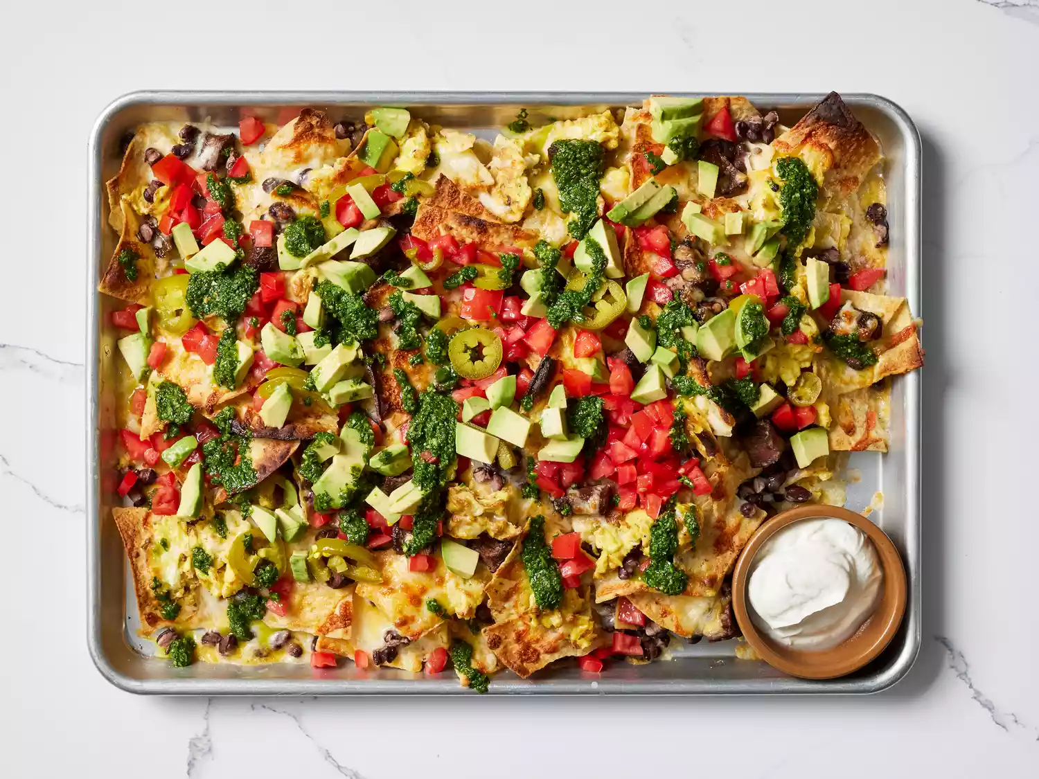 Breakfast Nachos garnished with Chimichurri and picked jalapeÃ±os on a baking sheet with a side of sour cream 
