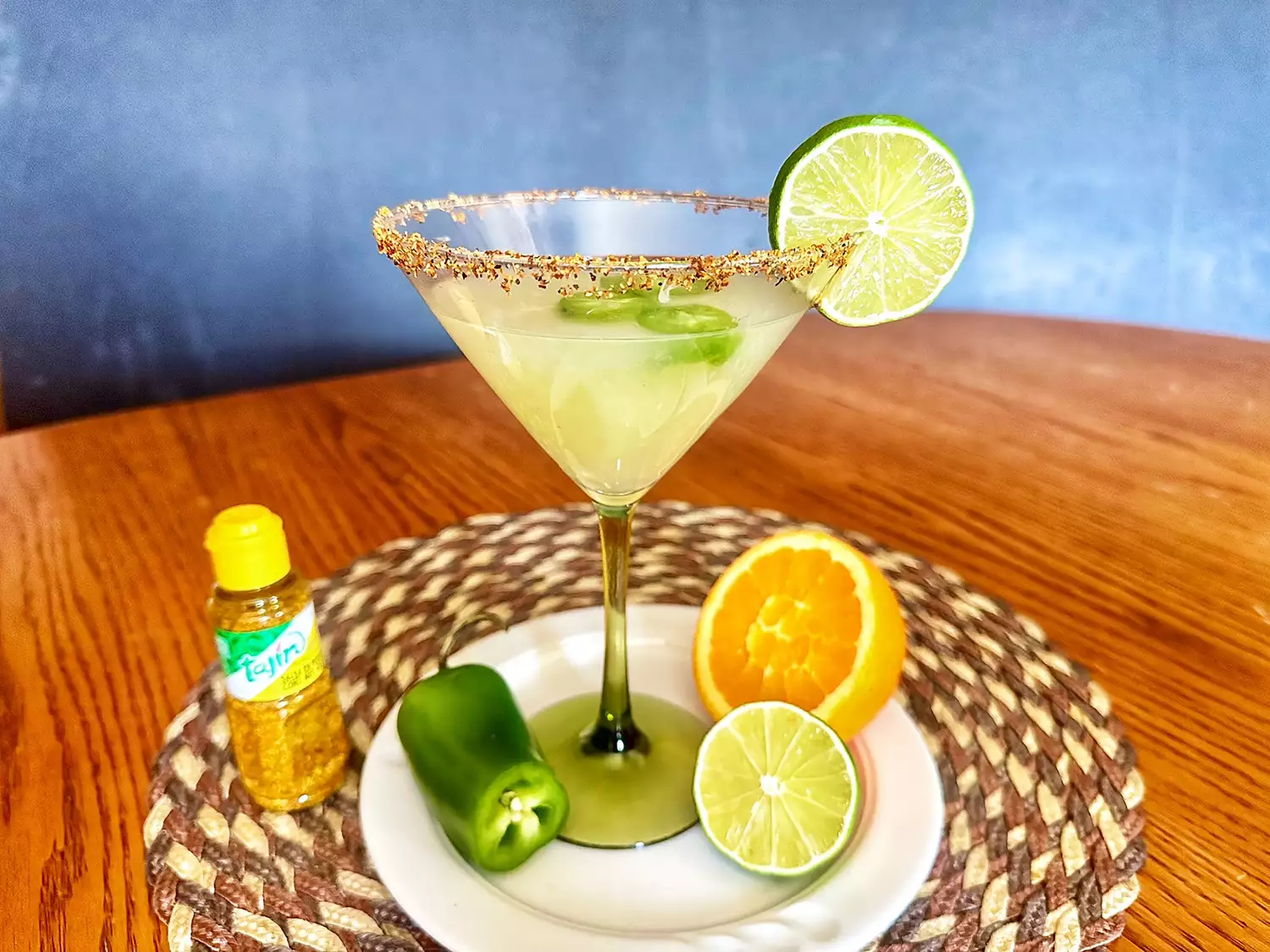 Margarita on ice with tajin seasoning on woven mat on a wooden table with jalapeno, half lime, and half orange in view.