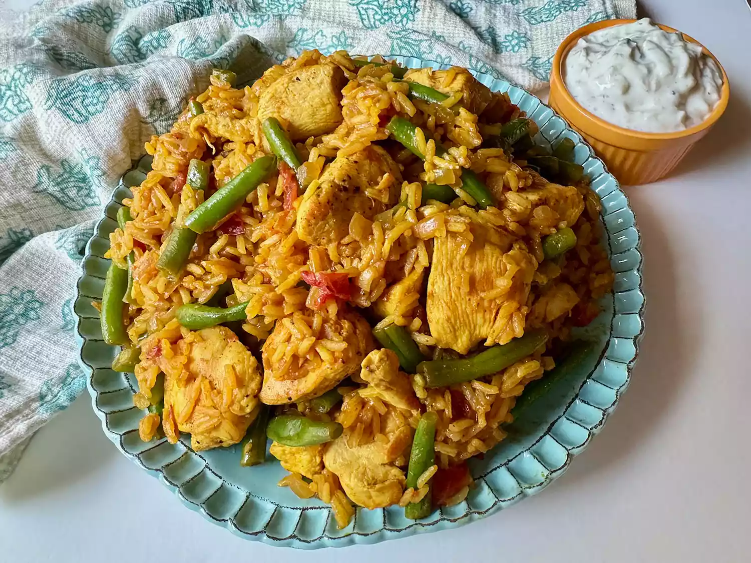 Overhead view of turmeric chicken and rice with green beans and tomatoes on aqua plate with indian print aqua towel and yellow ramekin of raita