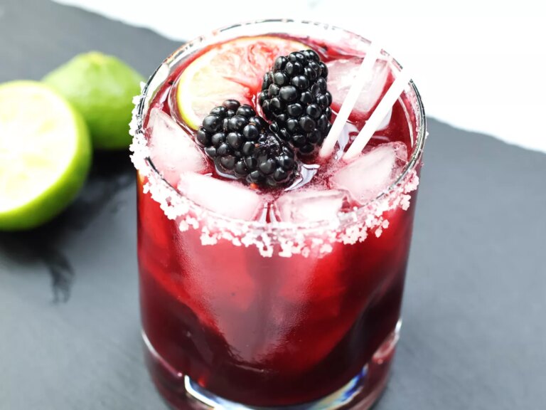 Clear rocks glass of blackberry margarita with salted rim, topped with 2 blackberries and lime slice