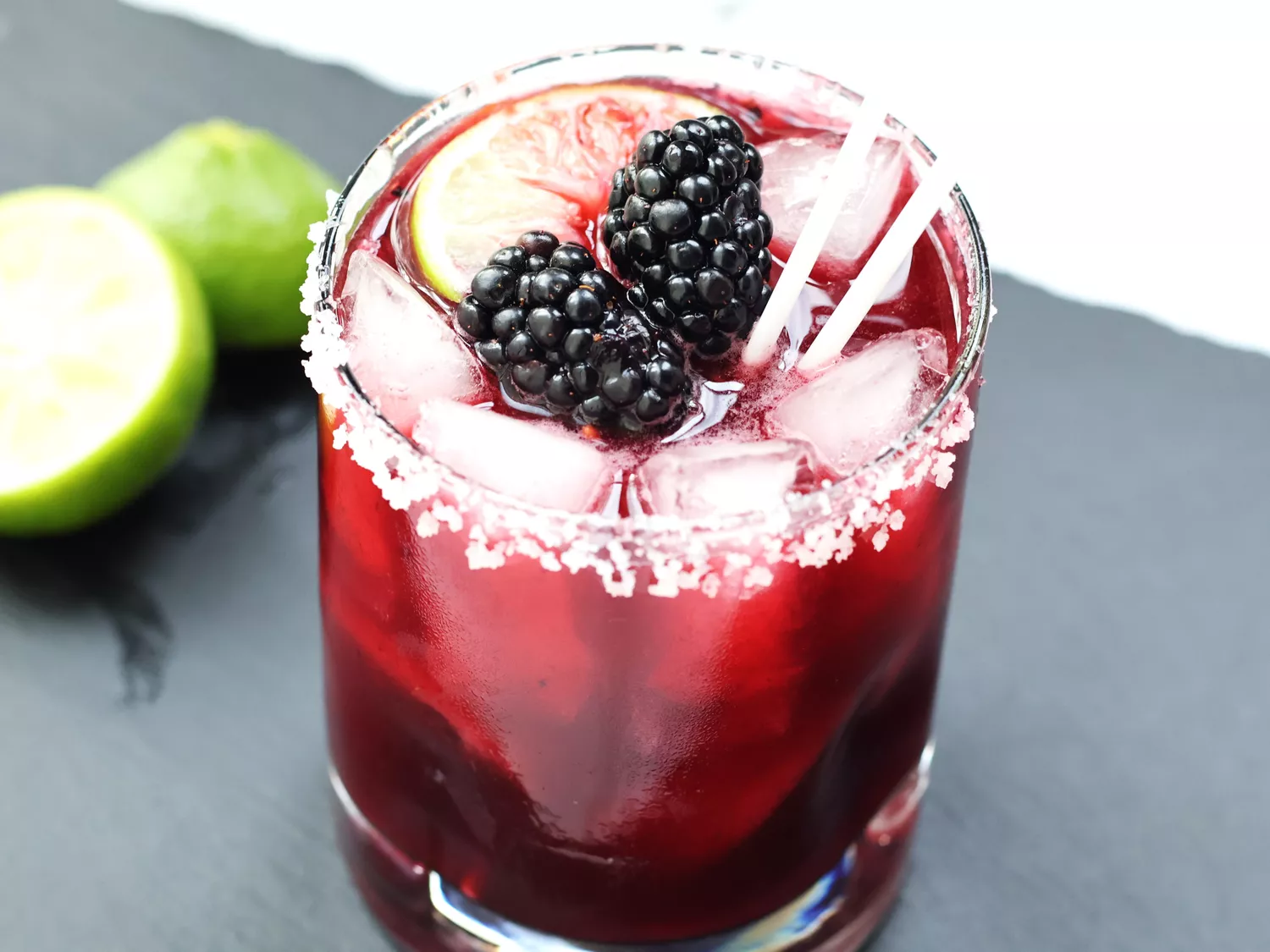 Clear rocks glass of blackberry margarita with salted rim, topped with 2 blackberries and lime slice