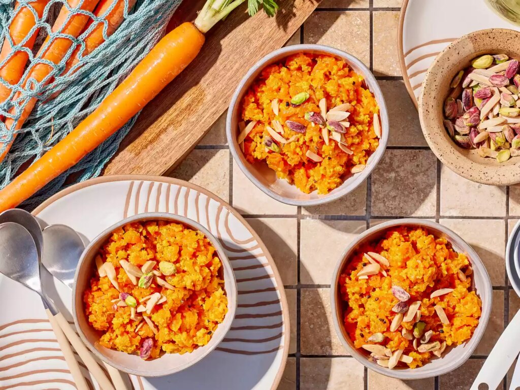 Carrot halwa in bowl garnished with nuts and cardamom next to plates, utensils and carrots on a wooden cutting board