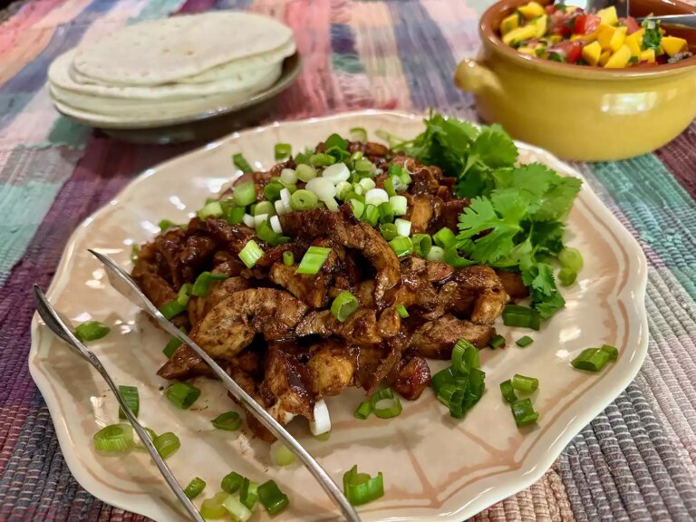 Sliced adobo chicken thighs on plate with cilantro and green onion and mango salsa and tortillas in background