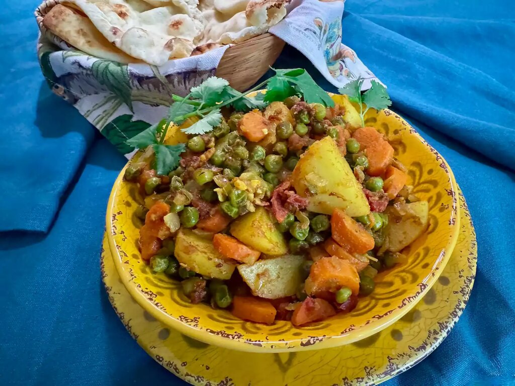 Yellow bowl of mixed vegetable curry with basket of naan on blue background